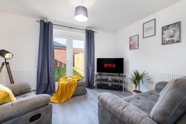 A living room with grey sofas, a yellow throw blanket, a TV displaying 'NETFLIX', and a plant. - The Holt (Lincoln)