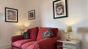 Un sofá rojo con dos almohadones decorativos, una mesita de noche con una lámpara y tres cuadros enmarcados en la pared.