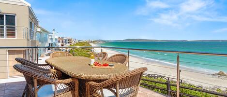 Una mesa y sillas de mimbre en un balcón con vistas a una playa y una vista despejada del océano y colinas distantes.