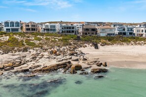 A coastal area with a sandy beach, rocky shoreline, and a row of modern houses with a staircase leading to a higher elevation. - Tenos On Calypso 16 (Langebaan)