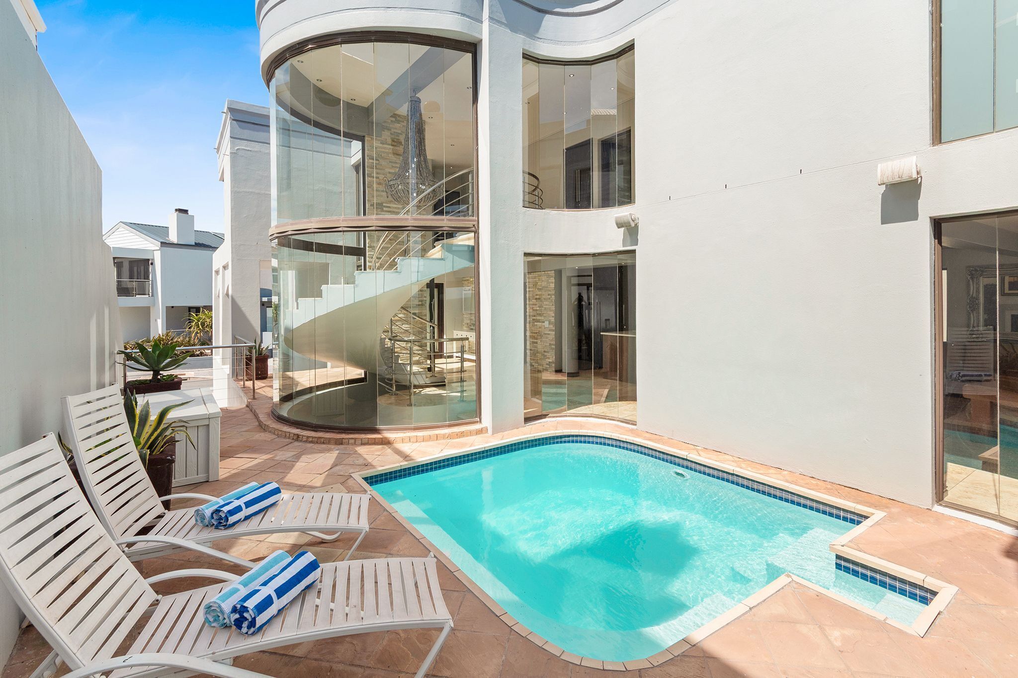 A modern building with a swimming pool, white lounge chairs, and a spiral staircase.