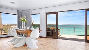 Een moderne eetruimte met een houten tafel, witte stoelen en een bloevas. Er zijn grote ramen met uitzicht op de zee en een balkon met buitenmeubilair.