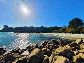 A rocky shoreline with clear blue water reflecting sunlight, a sandy beach, and a forested hill in the background.