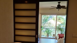 Une salle de bain avec une baignoire, une douche et des toilettes. Une porte coulissante donne accès à un balcon équipé d’un lit et d’un ventilateur de plafond.