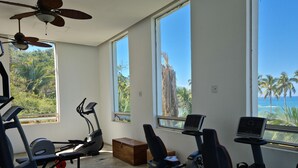 Une salle de sport équipée de matériel d’exercice, dotée de grandes fenêtres et offrant une vue sur les palmiers et l’océan.
