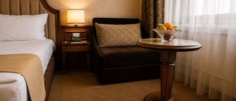 Habitación de hotel con una cama, una mesita de noche con lámpara, un sillón pequeño y una mesa redonda con fruta.