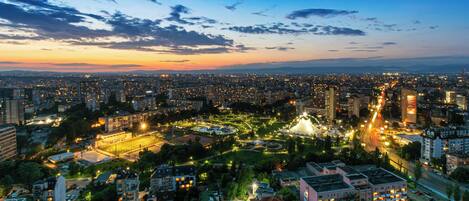 A cityscape at dusk with illuminated buildings and a park with a fountain.