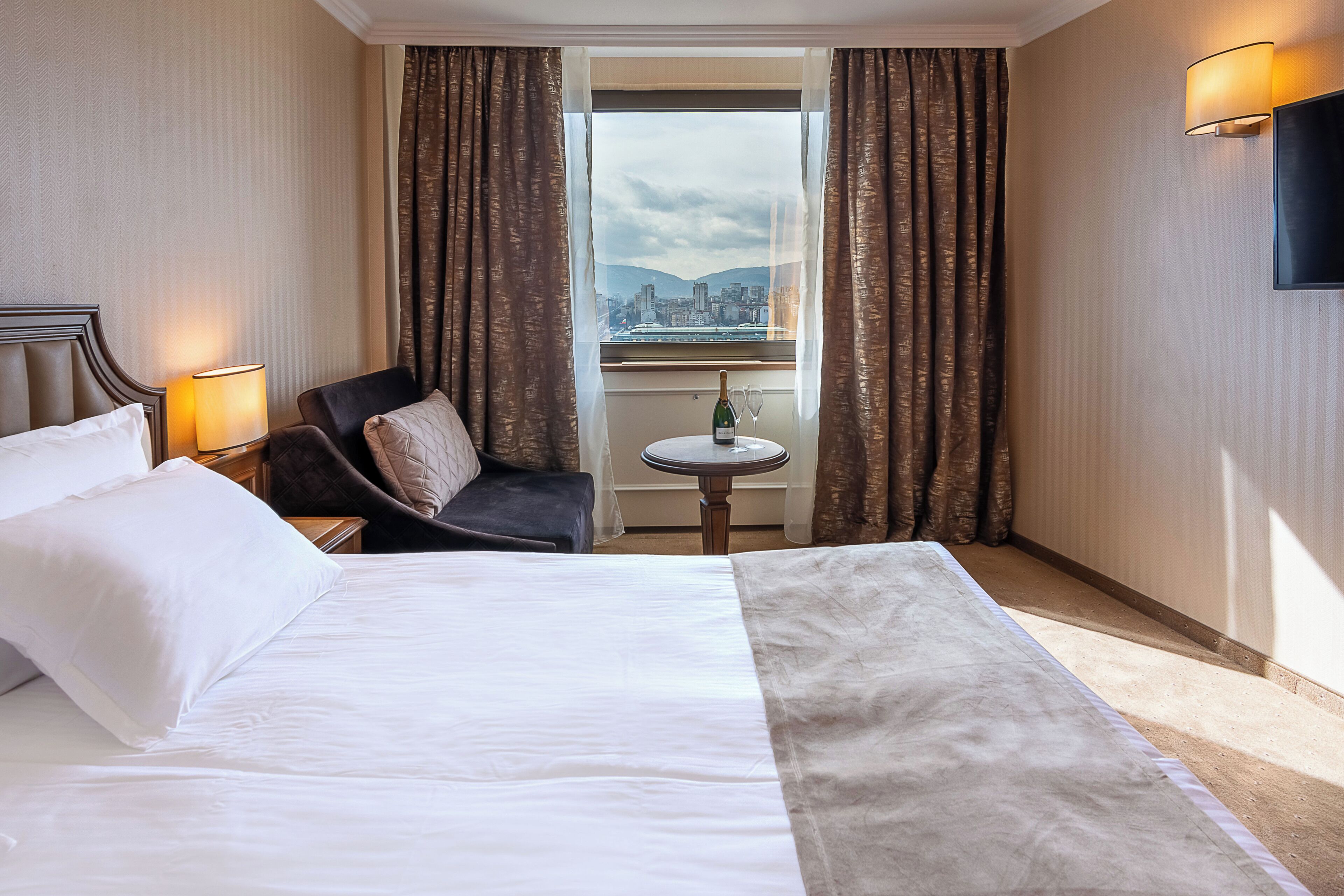 A hotel room with a bed, a chair, a small table with a bottle and glasses, and a view of a cityscape through a window.