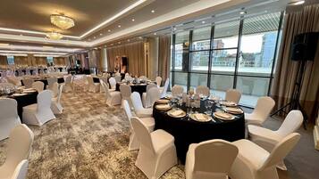 Un salón de banquetes con mesas redondas dispuestas para cenar, sillas blancas, candelabros y ventanas amplias con vista a la ciudad.