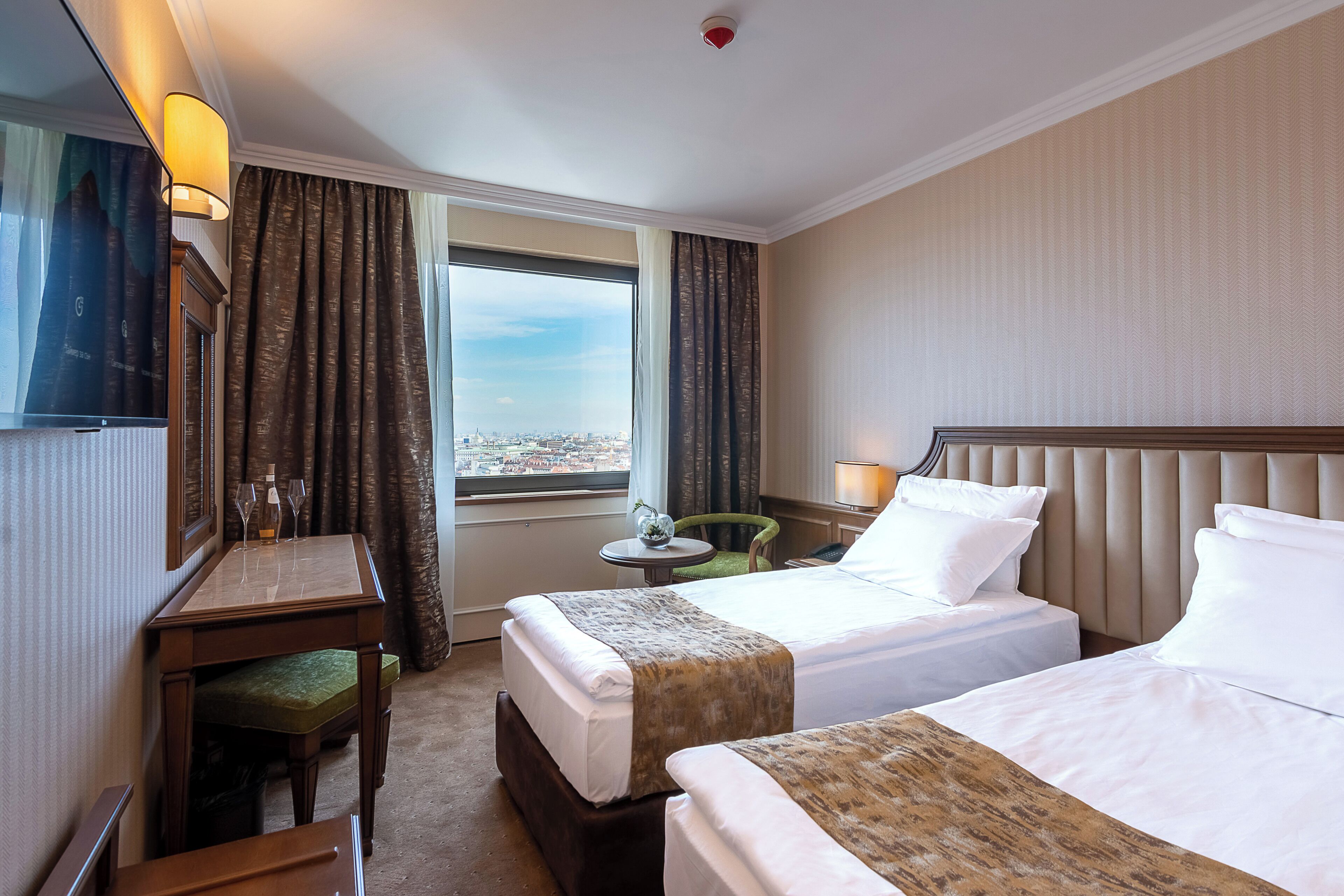 A hotel room with two beds, a desk, a chair, a small table, and a view of the city.