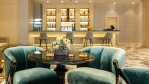 Una zona de bar con mostrador y estanterías llenas de botellas, dos sillones turquesa y una mesa con un centro de mesa floral.