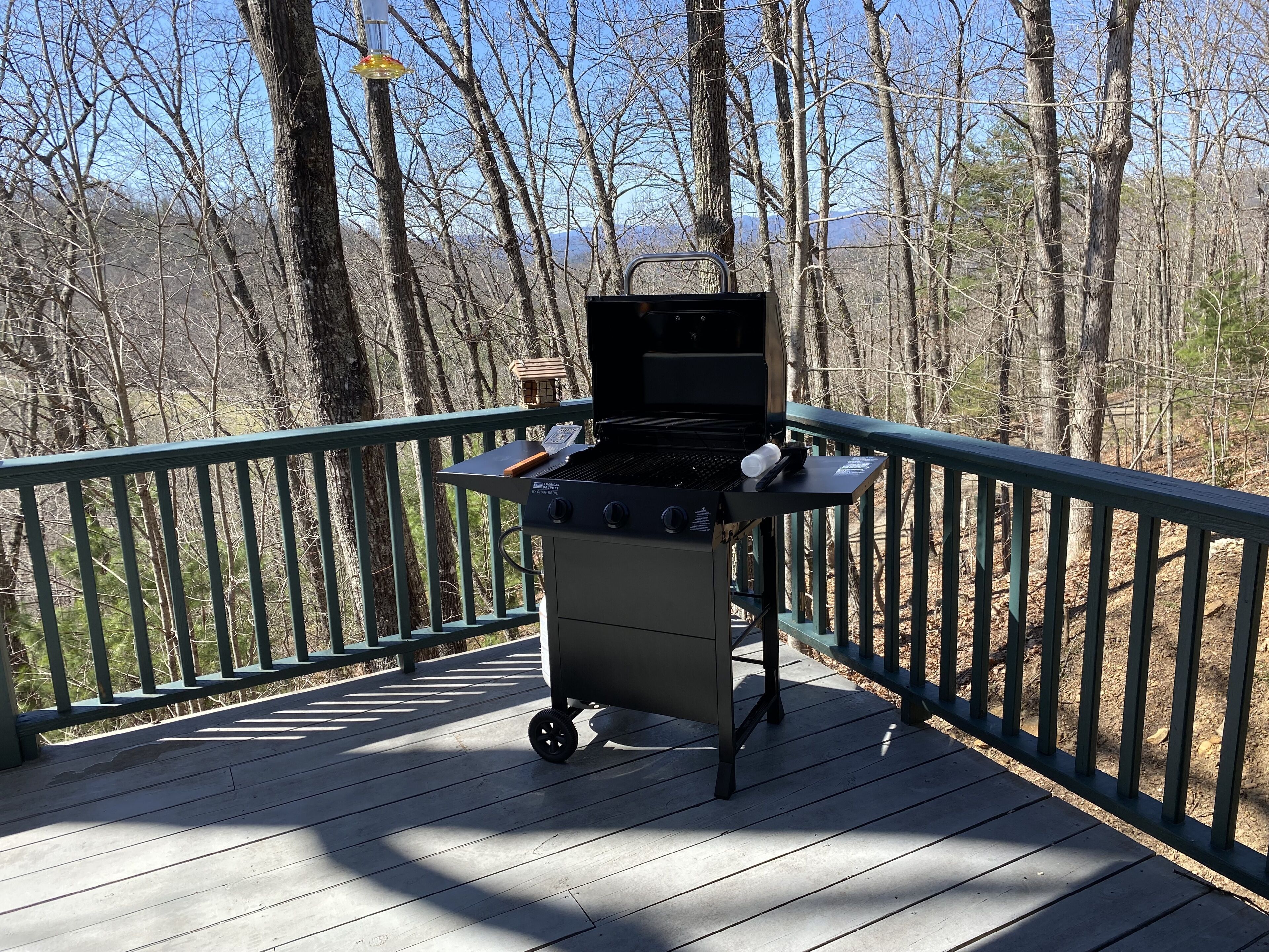 A portable grill on a wooden deck with a railing, surrounded by trees.