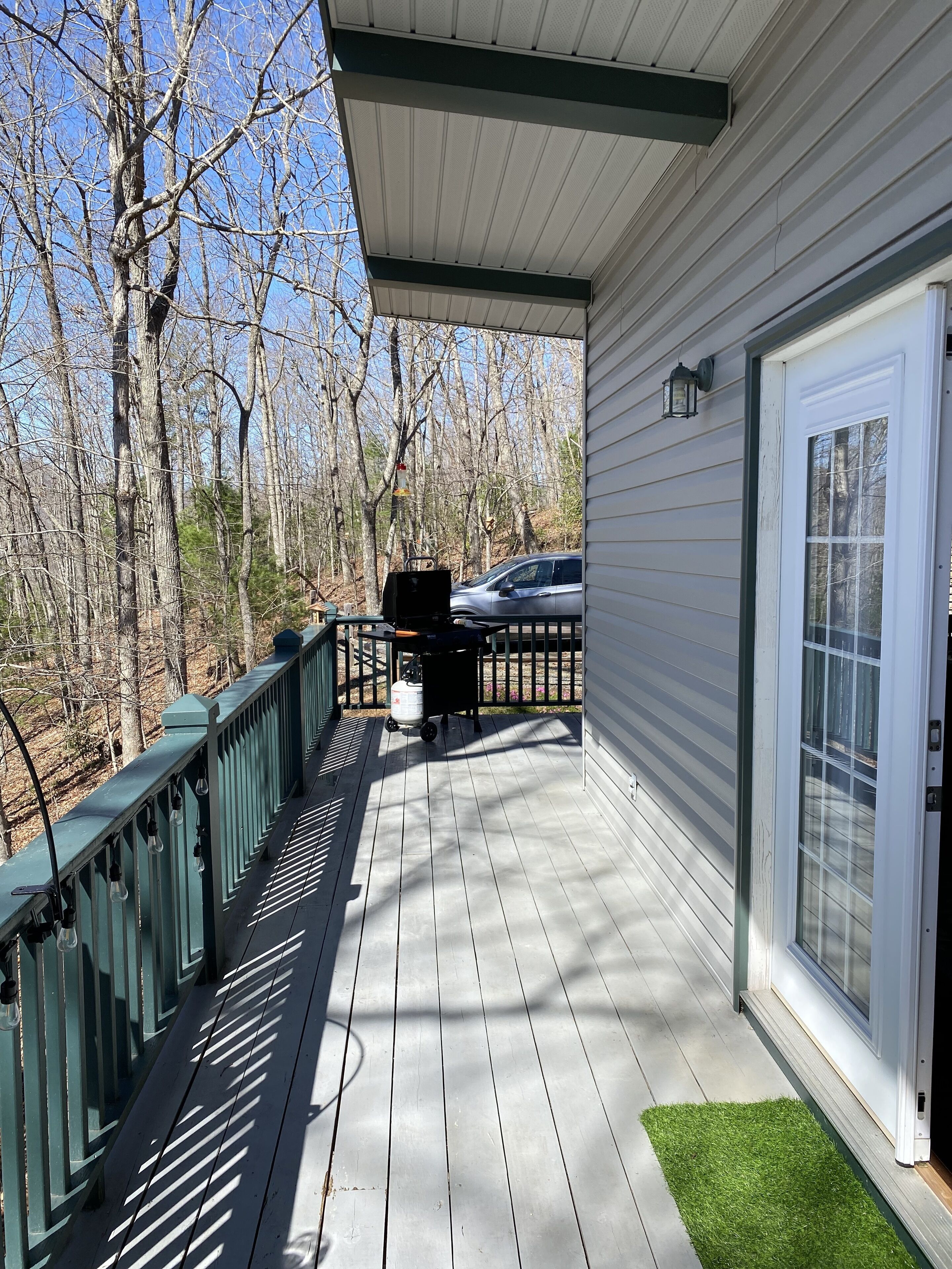 A covered porch with a grill, a car parked in the background, and a view of a wooded area.