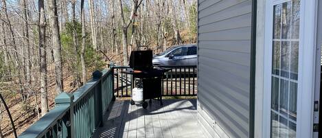 A covered porch with a grill, a car parked in the background, and a view of a wooded area.