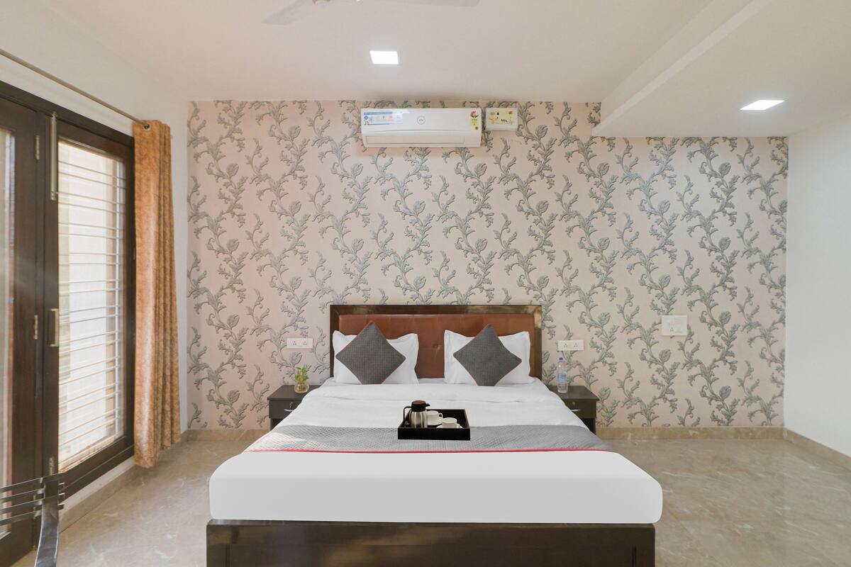 A hotel room with a bed, a nightstand, an air conditioning unit, and a window with blinds.