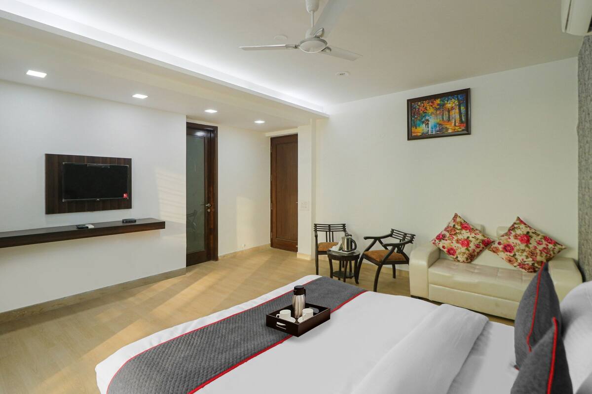 A hotel room with a bed, a TV mounted on the wall, a ceiling fan, a small table with chairs, and a sofa with floral cushions.