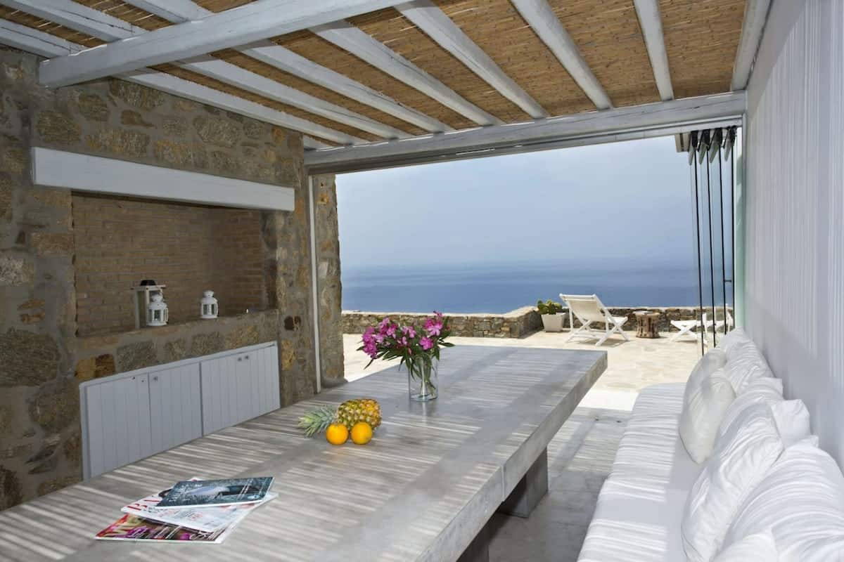 Een kamer met stenen muren, een houten plafond, een witte bank, een tafel met fruit en een vaas, en uitzicht op zee.