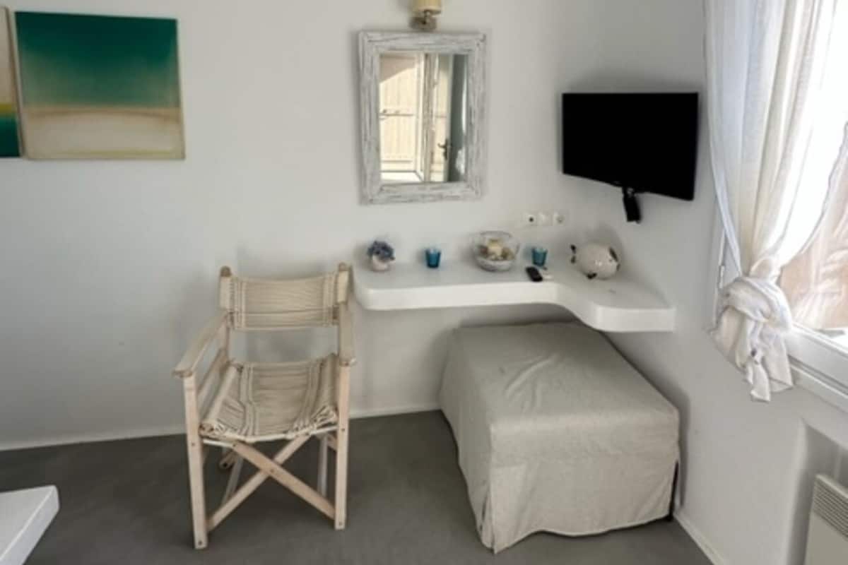 Een kamer met een wit plafond, een houten stoel, een klein bankje, een witte muur met een spiegel, een plank met decoratieve objecten, en een aan de muur bevestigde televisie.