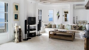 A spacious living room with a large TV, a wooden coffee table, and a sofa. There are framed pictures on the wall and a view of the sea through the windows. - White Rock Suites & Villas (Mykonos)