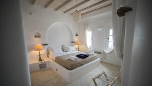 A bedroom with a large bed, two bedside lamps, a hanging light fixture, a small table, and a chair. - White Rock Suites & Villas (Mykonos)