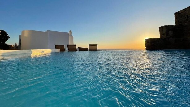 A pool with clear blue water, a sunset in the background, and a stone structure on the right. - White Rock Suites & Villas (Mykonos)