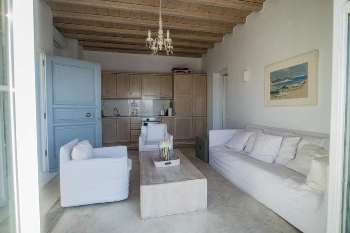 Een moderne woonkamer met een witte bank, een salontafel, en een eetgedeelte met houten kasten en een kroonluchter.