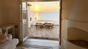 A room with a view of the sea at sunset, a sofa, a table, and a small fireplace. - White Rock Suites & Villas (Mykonos)