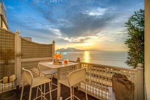 A balcony with a table and chairs, overlooking a body of water and mountains during sunset. - Honeymooners nest in Amalfi coast  (Marciano)