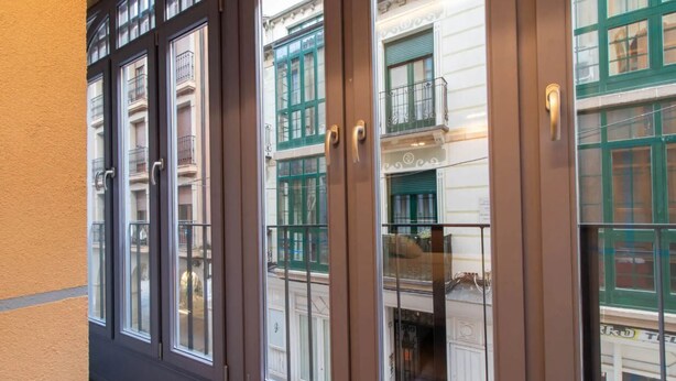 A brown-framed window with a glass door, reflecting a multi-story building with green window frames and balconies. - Hotel Cortabitarte (Soria)