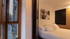 A hotel room with a bed, a painting on the wall, and a window overlooking a street. - Hotel Cortabitarte (Soria)