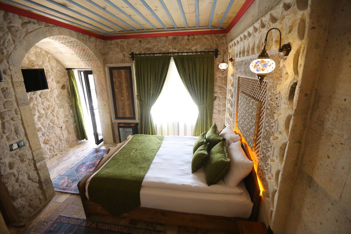 Quarto com parede de pedra, cama com roupa de cama verde, tapete estampado e luminária fixada na parede.