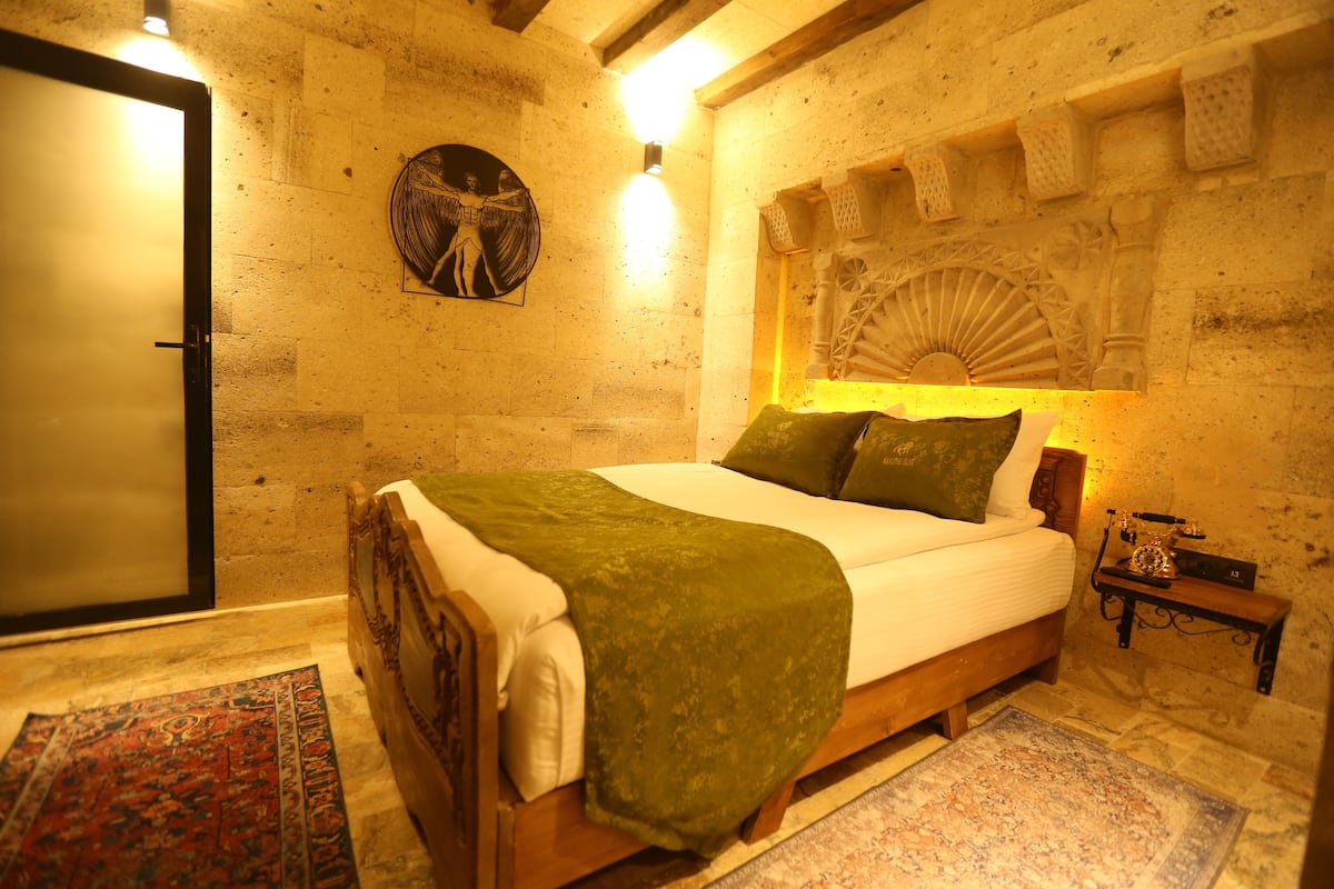Quarto com cama de madeira, manta verde, mesa de cabeceira com abajur e ventilador fixado na parede.