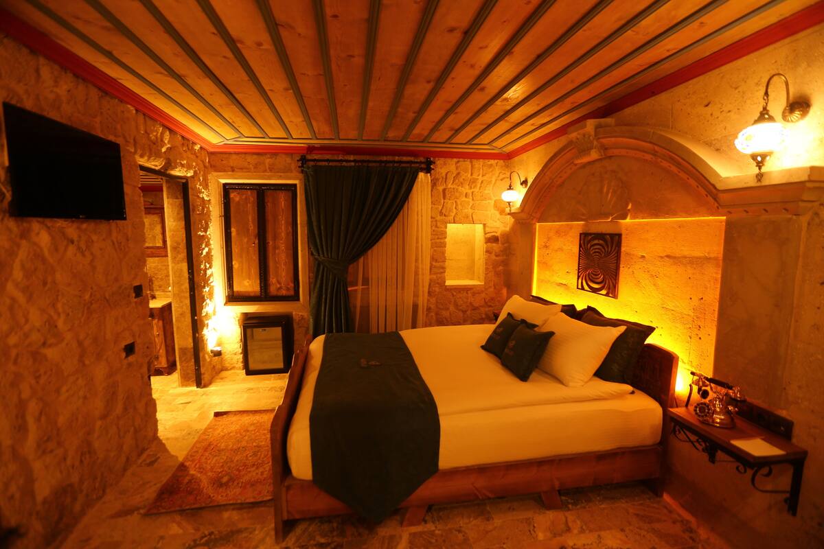 Quarto aconchegante com uma cama grande, paredes de pedra, vigas de madeira, lareira de pedra e uma televisão montada.