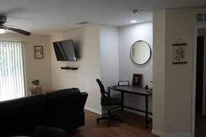 A living room with a black leather sofa, a flat-screen TV mounted on the wall, a round mirror, and a desk with a calendar. - Peaceful Lehigh Duplex, 200+ 5 star, Clean & Comfortable Stays (Lehigh Acres)
