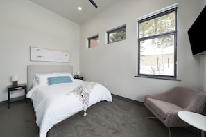 Quarto moderno com cama, mesas de cabeceira, uma cadeira e uma televisão.