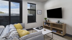 A modern living room with a grey sofa, yellow pillow, and a coffee table. A flat-screen TV is mounted on the wall, and there are books and decorative items on a wooden shelf.
