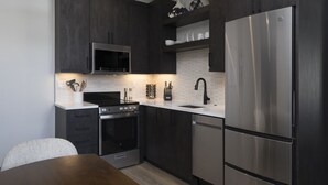 A modern kitchen with dark cabinetry, stainless steel appliances, a white countertop, and a dining area with a wooden table.