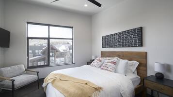 A modern hotel room with a large bed, a view of a building through a window, a chair, and a desk with a lamp.