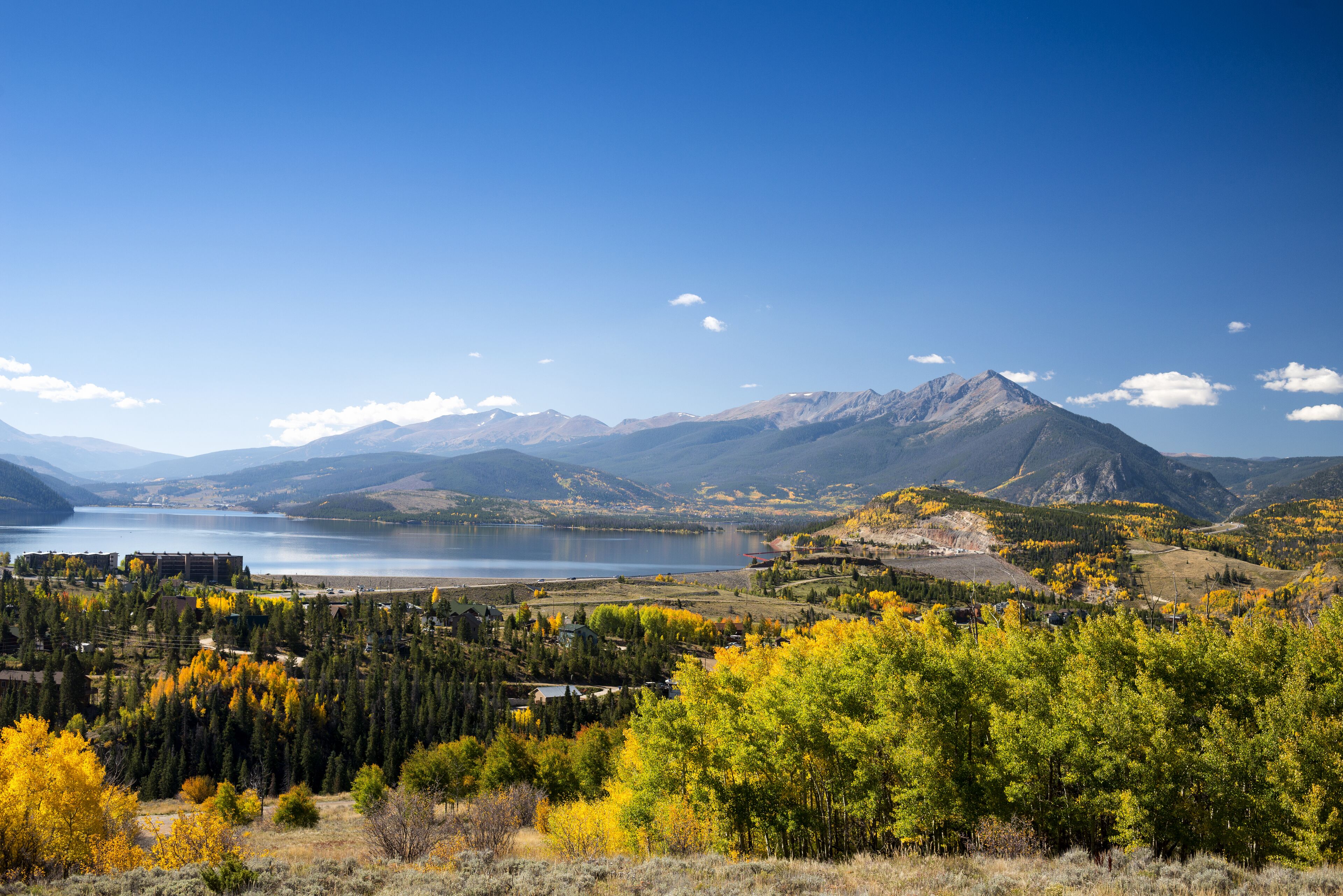 Paisaje montañoso con un lago, follaje otoñal y un pequeño pueblo.