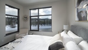 Un dormitorio con una cama grande, dos ventanas, un ventilador de techo, una mesita de noche con una lámpara y vistas a un paisaje nevado.
