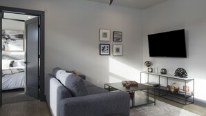 Una sala de estar moderna con un sofá gris, una mesa de centro y un televisor montado en la pared. Se observan cuadros enmarcados en la pared y una estantería con objetos decorativos.