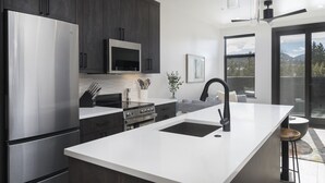 Une cuisine moderne avec des meubles de couleur sombre, des appareils en acier inoxydable, un grand îlot central doté d’un robinet noir et une vue sur les montagnes grâce à de grandes fenêtres.