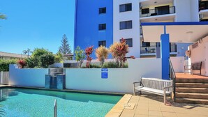 Zona de piscina amb una piscina de blau clar, una boscadeta de fusta i una escala que mena a un edifici amb parets blaves i blanques.