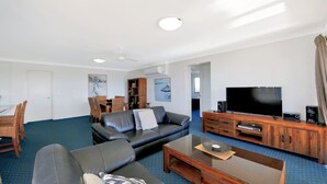 A modern living room with a black leather sofa, a wooden entertainment unit with a TV, and a dining area with wicker chairs.