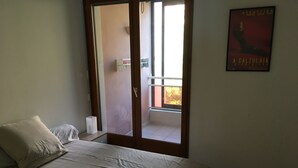 Un dormitorio con cama, almohada, mesita de noche, ventana y puerta que da a un balcón.