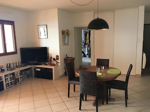 A living room with a TV, a dining table with chairs, and a kitchen area visible through an open doorway.