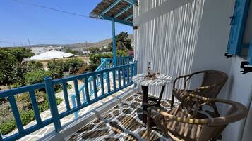 A balcony with a table and chairs, a view of the countryside, and a white building with blue accents.
