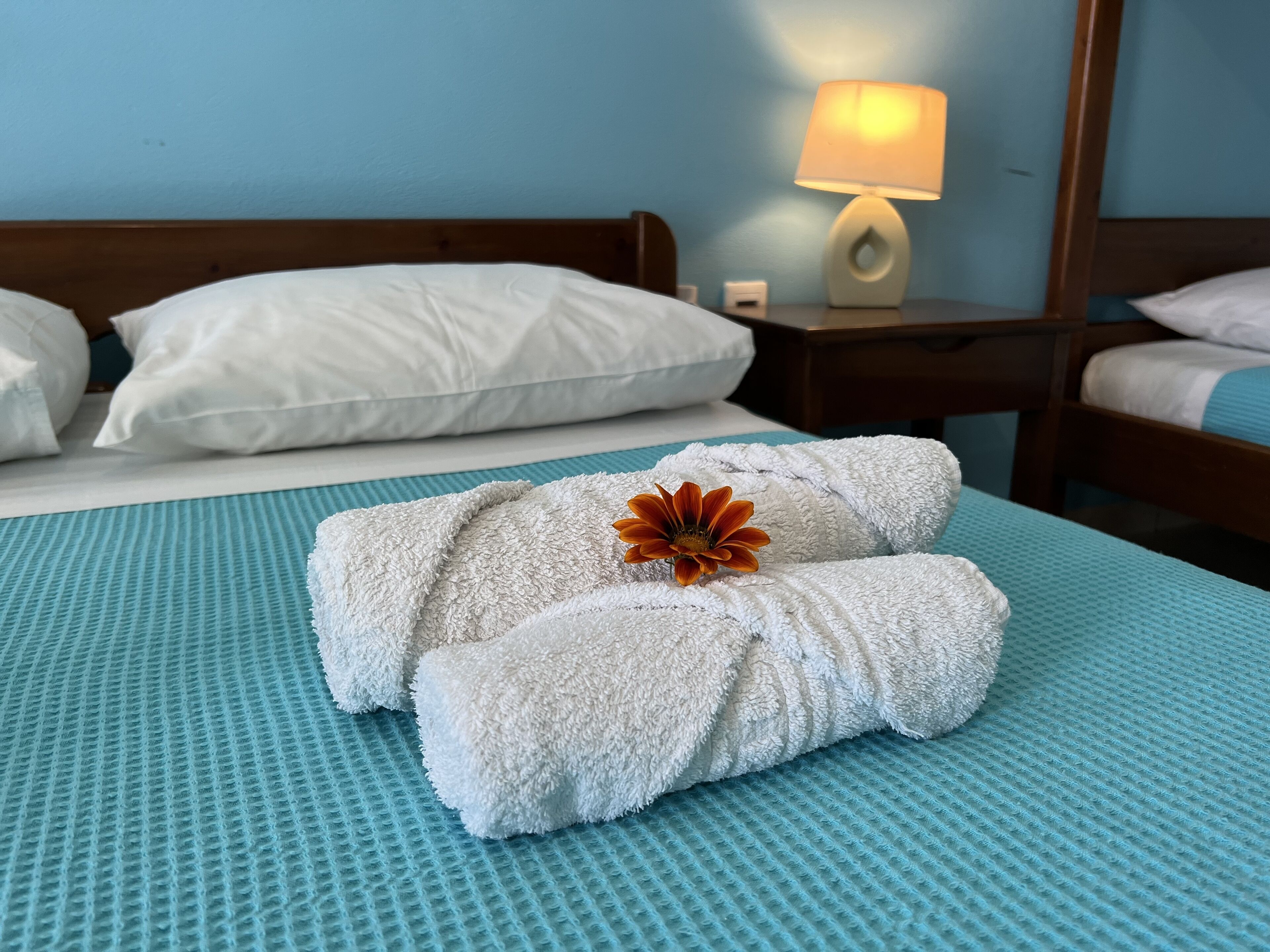 A neatly made bed with white pillows and a rolled white towel with a flower on it.