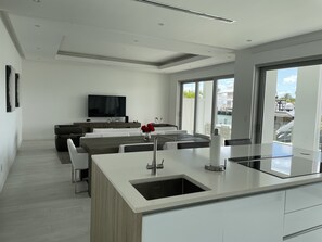 Eine moderne Küche mit Kochinsel, Essbereich inklusive Sofa und Stühlen, ein flachbildfernseher an der Wand und große Fenster mit Blick ins Freie.