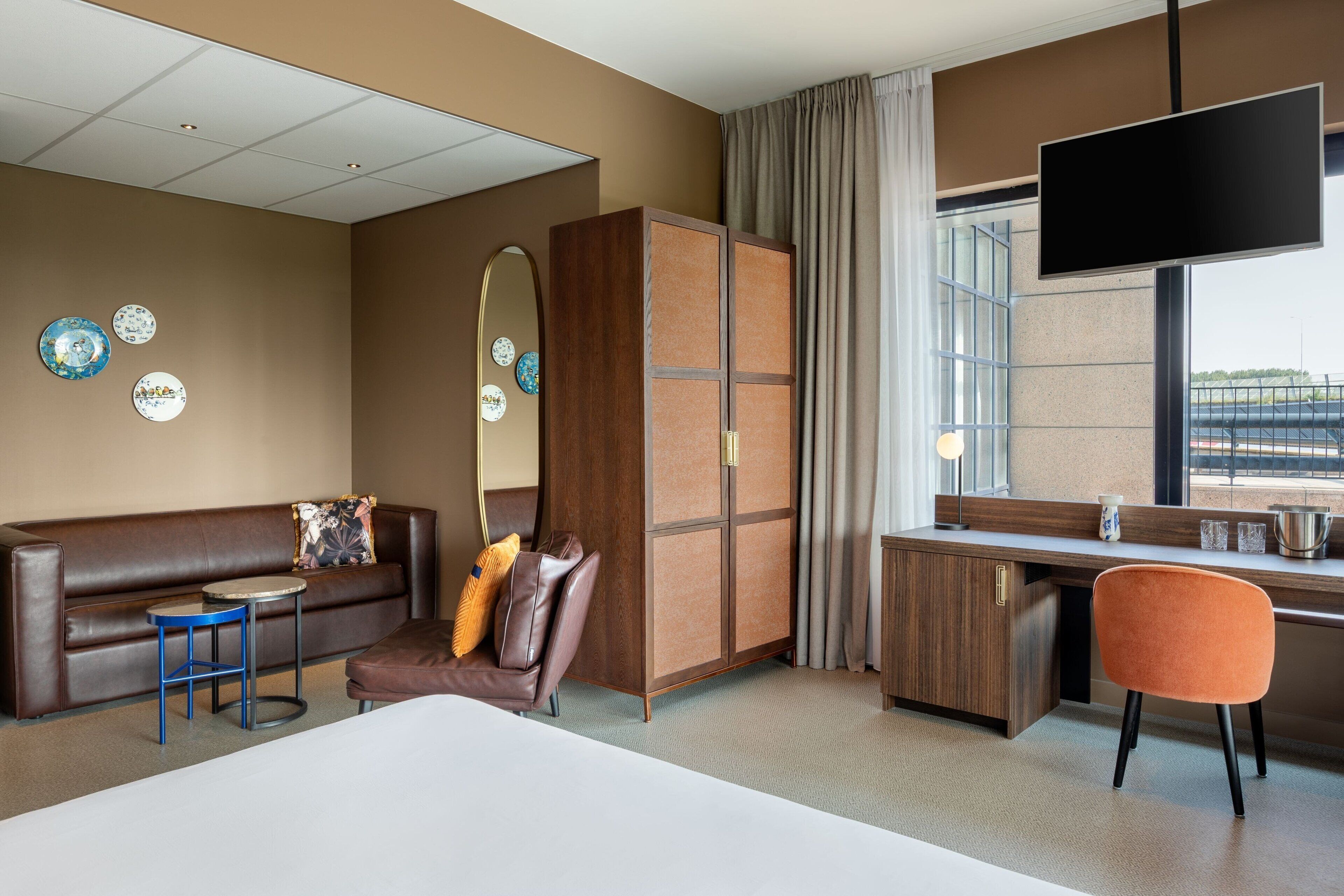 Una habitación de hotel con una cama, un sofá de cuero marrón, una mesita redonda pequeña, un escritorio con una silla, un armario, un espejo grande y un televisor montado en la pared.